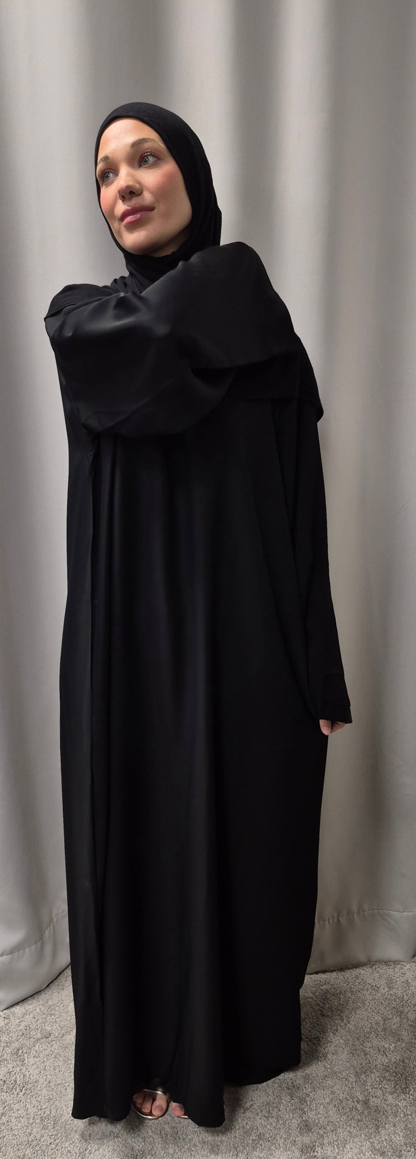A woman models the Shop Layla Closed Abaya - Classic Black Everyday Elegance from Bihi Boutique, standing before a light gray curtain with a soft expression and one arm bent across her body.