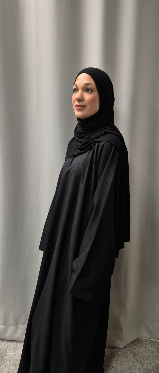 A woman in the Elegant Linen Zamzam Black Abaya by Bihi Boutique (Size 54) stands before light curtains, gazing upward with a gentle smile. Shop Now!.