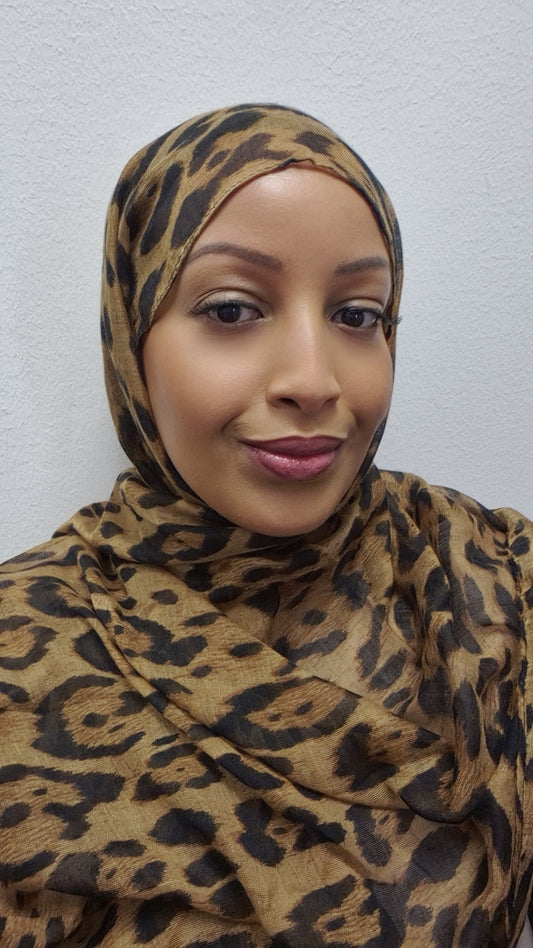 A woman models the Stylish Cheetah Hijab from Bihi Boutique, smiling softly at the camera while standing against a plain white textured wall.