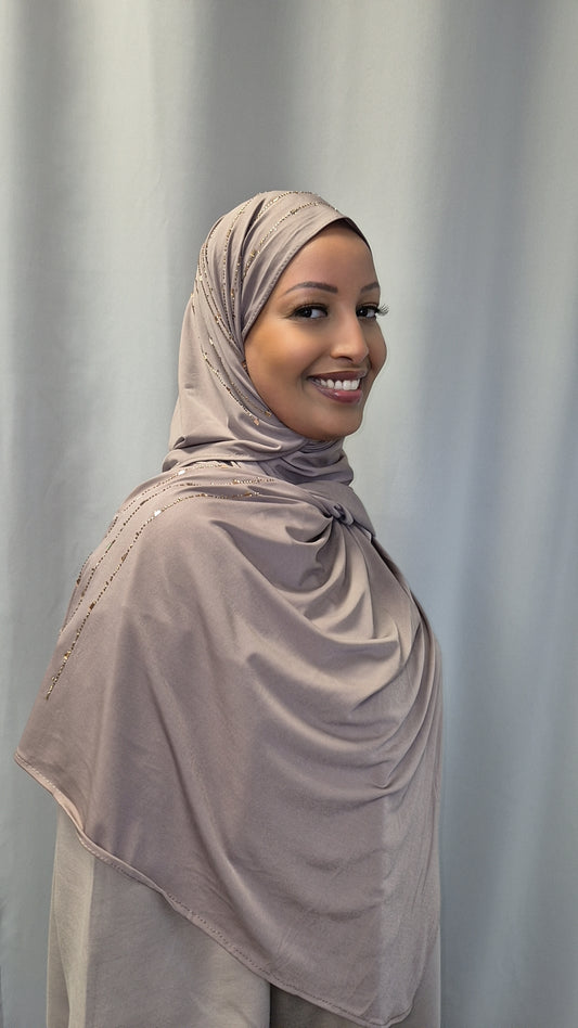 A woman in the Shop Premium Nude Jersey hijab by Bihi Boutique, featuring a trendy and stylish light beige design, stands smiling in side profile before a plain light gray curtain.