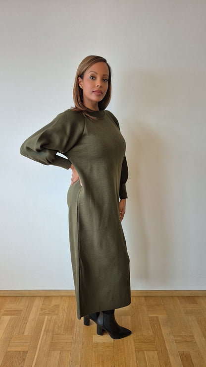 A woman with light brown hair poses indoors by a white wall, wearing the Olive Green Sweater Dress from Bihi Boutique with black heeled boots, one hand on her hip, looking slightly to the side.