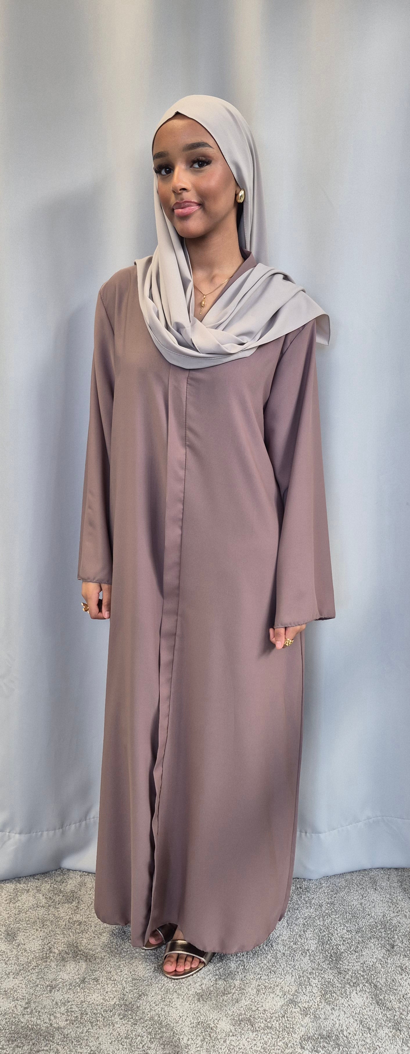 A woman models the Islene Dress by Bihi Boutique (fits 160-165 cm, taupe), paired with a light gray hijab, standing on a gray carpet before a light gray curtain and smiling at the camera.