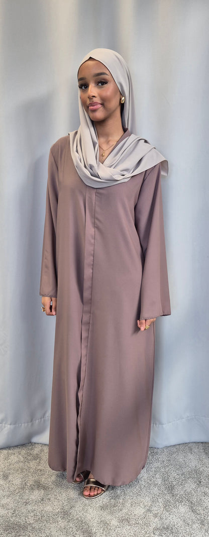 A woman models the Islene Dress by Bihi Boutique (fits 160-165 cm, taupe), paired with a light gray hijab, standing on a gray carpet before a light gray curtain and smiling at the camera.