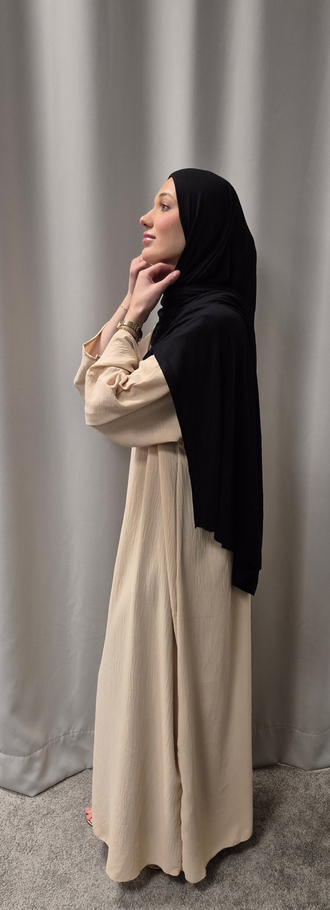 A woman in a Shop Gyllene Dress Size 60 by Bihi Boutique poses barefoot on a gray carpet, wearing a black hijab and touching her chin, standing in profile before a light gray curtain. Elegant & stylish fashion.