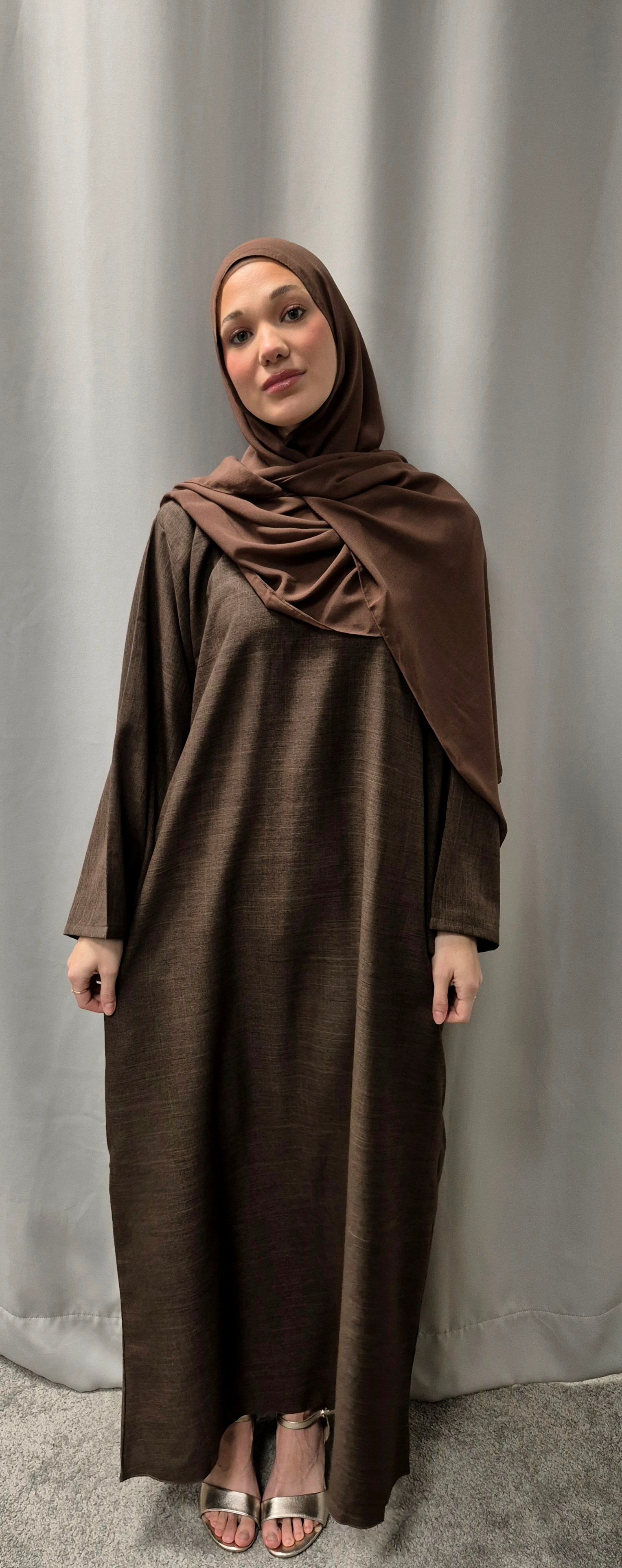 A woman models the Bihi Boutique Stylish Linen Zamzam Brown Abaya with Chiffon Hijab (Size 52), standing in front of a light gray curtain and looking at the camera. She pairs the outfit with open-toed sandals.