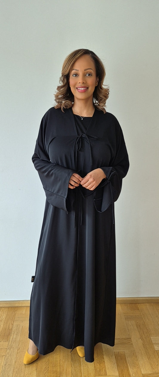 A woman smiles in the Bihi Boutique Ayan Black Nidah Dress with Chiffon Hijab (Size 60), standing on a wooden floor against a light-colored wall.