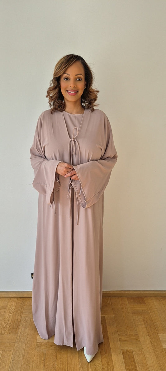 A woman with shoulder-length wavy hair smiles while wearing the Bihi Boutique Shop Ayan Nude Nidah Dress (includes Chiffon Hijab, Size 60) in light mauve, standing on a hardwood floor before a plain light wall.