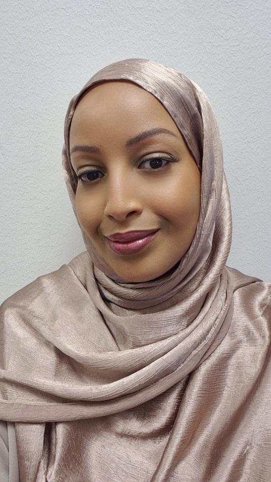 A woman models the Luxurious Satin Hijab from Bihi Boutique (173cm x 73cm) in shiny light beige, smiling softly at the camera against a simple textured light background.