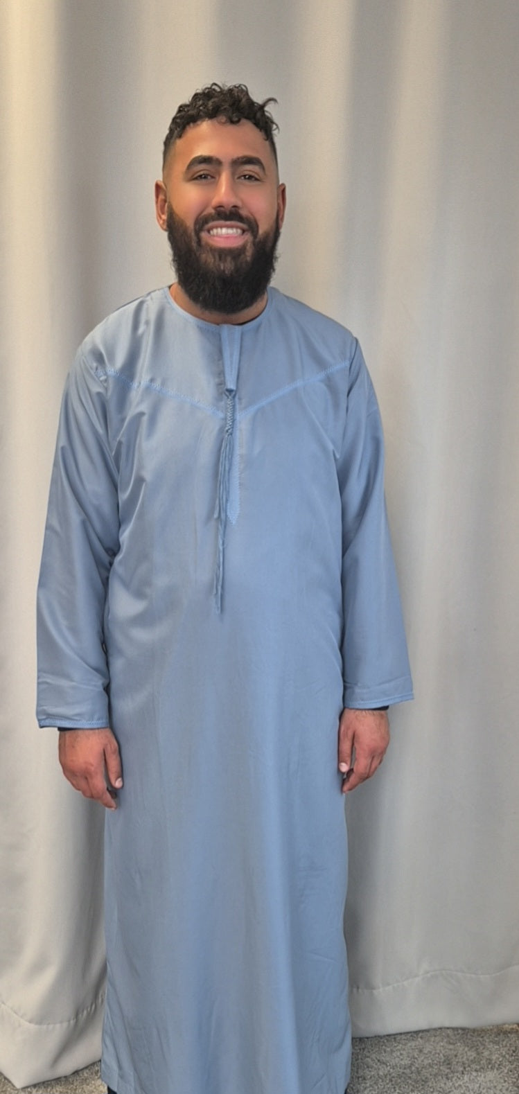 A man with dark hair and a beard smiles in front of a light curtain, wearing the Stylish Emirati Khamis from Bihi Boutique—luxury traditional wear perfect for all occasions.