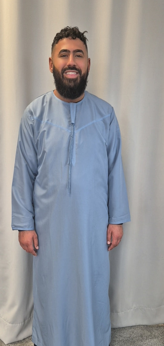 A man with dark hair and a beard smiles in front of a light curtain, wearing the Stylish Emirati Khamis from Bihi Boutique—luxury traditional wear perfect for all occasions.