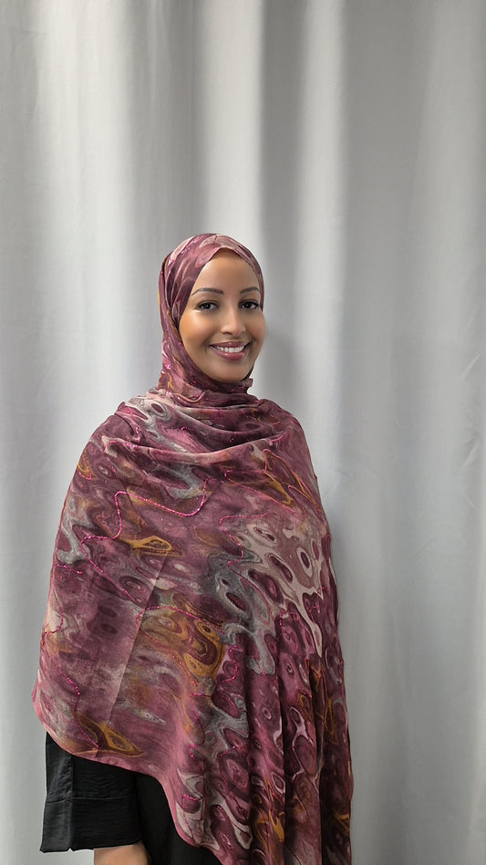 A woman smiles in front of a light gray curtain, wearing the Elegant Chiffon Hijab by Bihi Boutique—a lightweight, stylish maroon patterned hijab—paired with a black top.