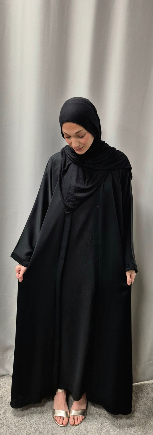 A woman models the Bihi Boutique Stylish Layla Open Abaya in Size 58, showcasing its elegant black design against a light grey curtain, as she gently holds the sides of the abaya while standing barefoot on a grey carpet.