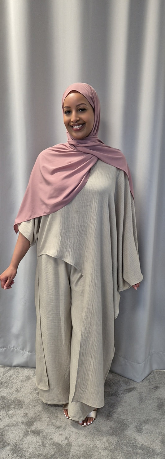 A smiling woman stands before a gray curtain, dressed in the Shop Luxurious 2-Piece Linen Set – One Size from Bihi Boutique, featuring a loose light beige fit with wide sleeves and pants, paired with a pink hijab over her shoulders.