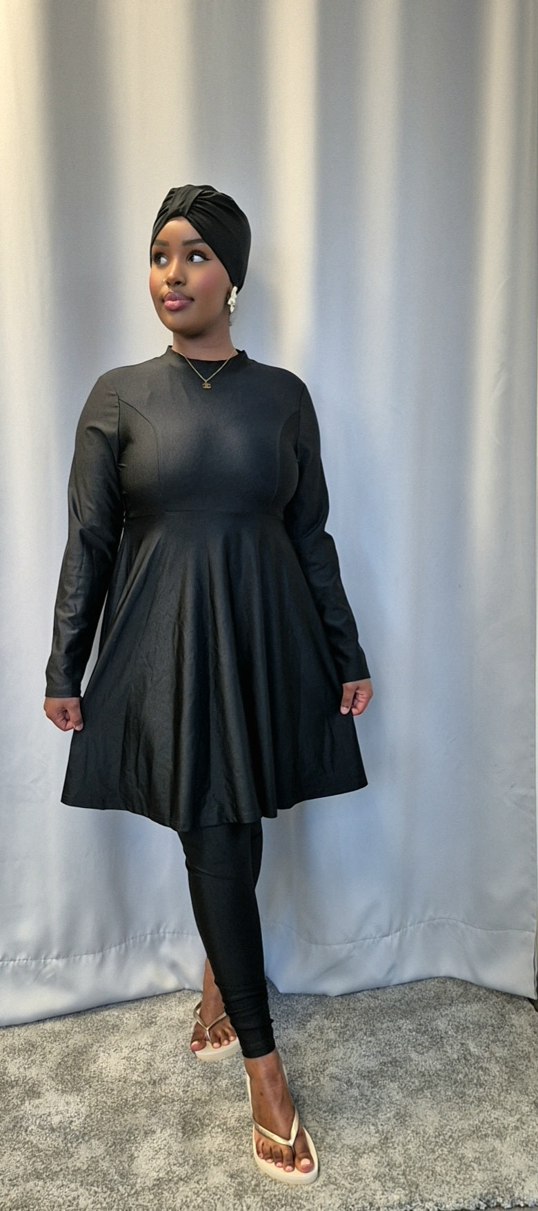 A woman models the Shop Stylish Black Burkini by Bihi Boutique, featuring a long-sleeve dress, leggings, and headwrap. She stands on a gray carpet in front of a light curtain, gazing slightly away with one hand by her side.