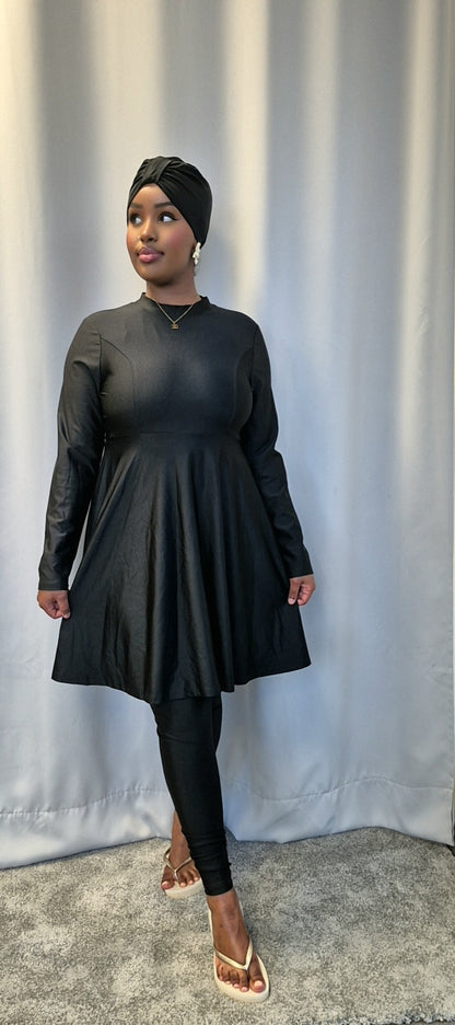 A woman models the Shop Stylish Black Burkini by Bihi Boutique, featuring a long-sleeve dress, leggings, and headwrap. She stands on a gray carpet in front of a light curtain, gazing slightly away with one hand by her side.