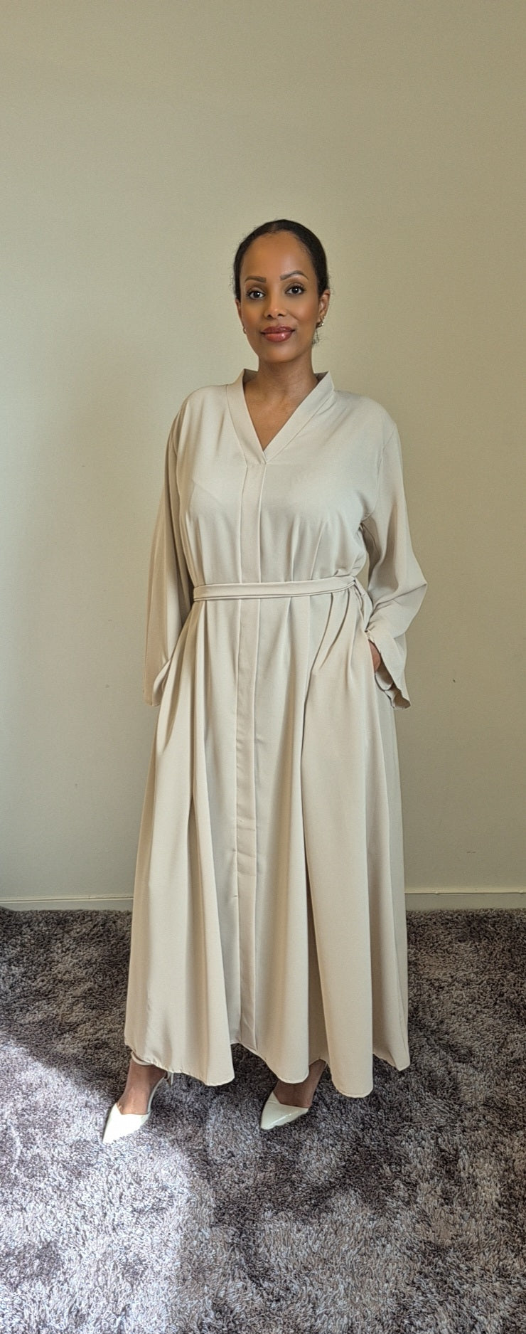 A woman stands indoors wearing the Shop Layina Black Dress by Bihi Boutique in L/XL, featuring long sleeves, a belted waist, and pockets. She looks at the camera against a plain light-colored wall, paired with white shoes.