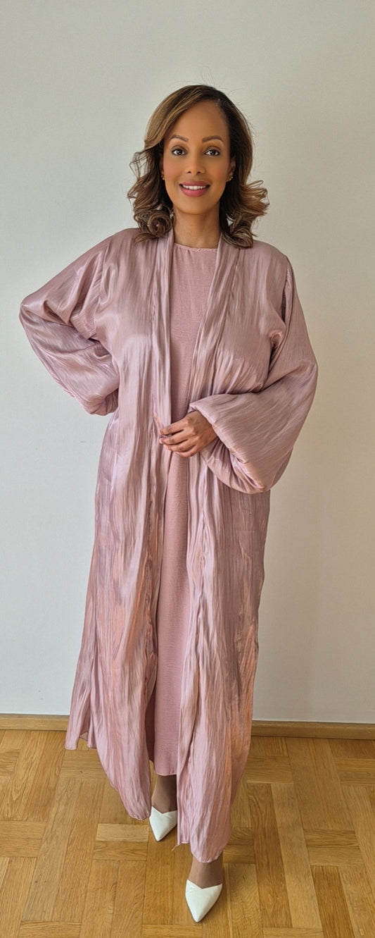 A woman stands on a wooden floor, smiling, in Bihi Boutique’s Elegant Pink Organza Slipdress with a matching chiffon hijab. She pairs the outfit with white pointed-toe heels, set against a plain white wall. Shop now!.