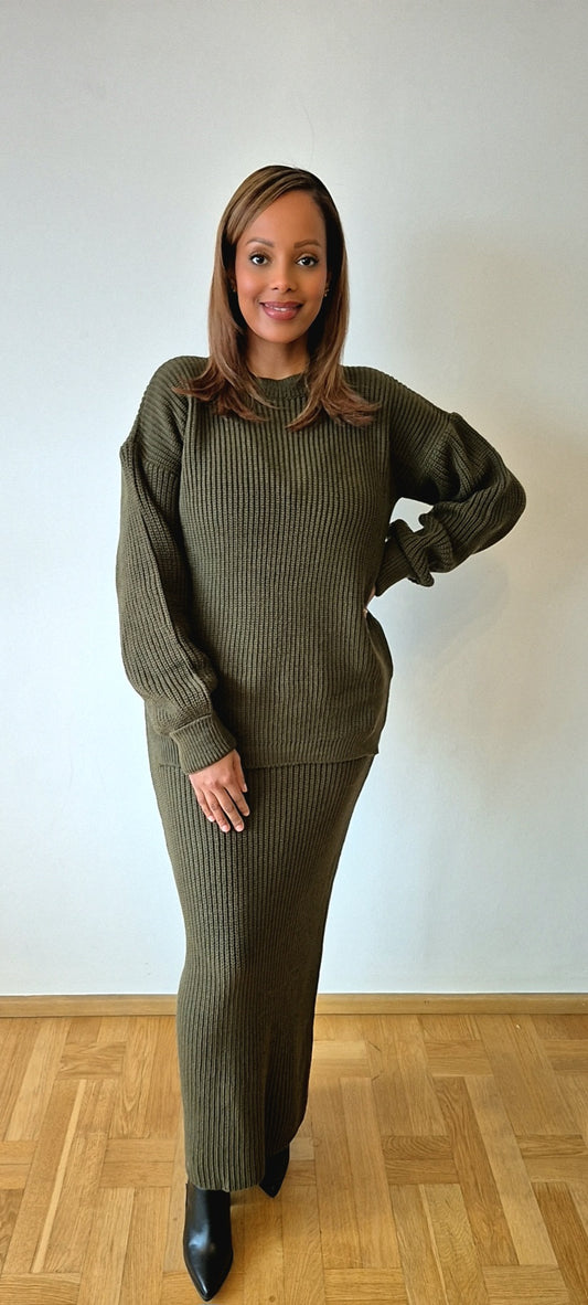 A woman stands indoors on a wooden floor, smiling with one hand on her hip, wearing the Chic Olive Green Sweater Skirt set from Bihi Boutique—a cozy wool blend ensemble in one size—paired with black pointed-toe boots.