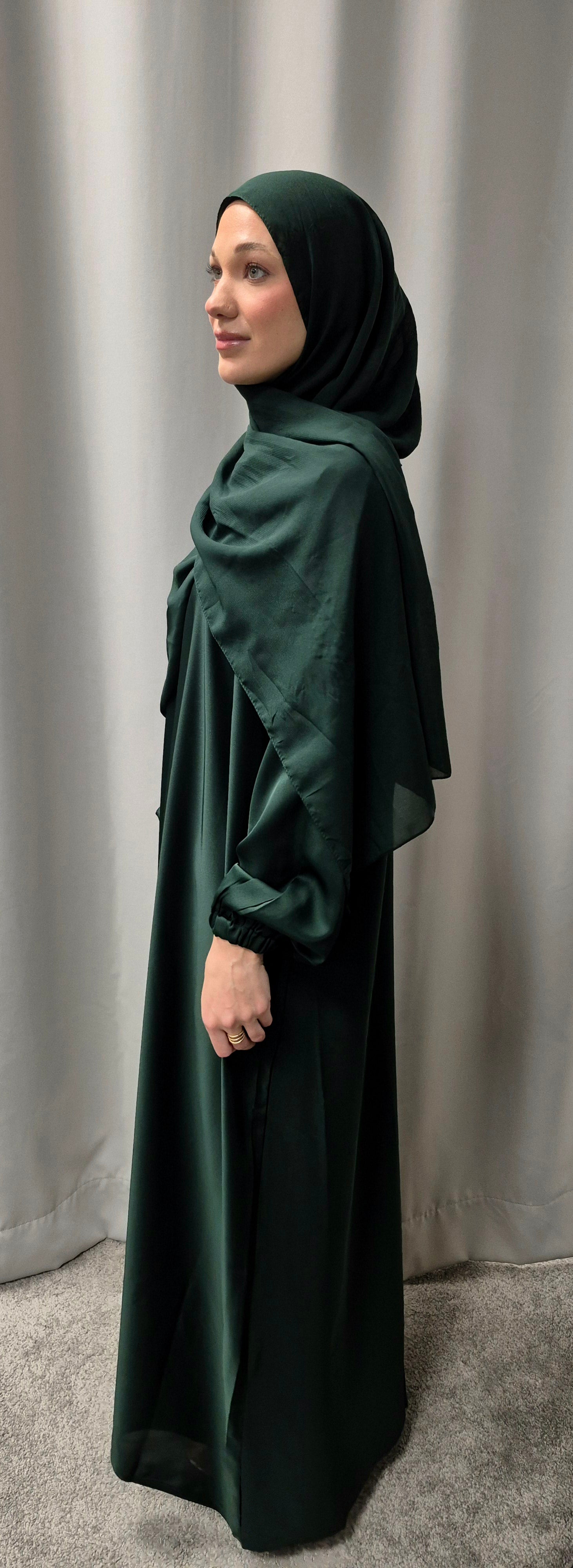 A woman wears the Shop Amina Abaya with Chiffon Hijab in Size 56 by Bihi Boutique, standing sideways against a light curtain backdrop.