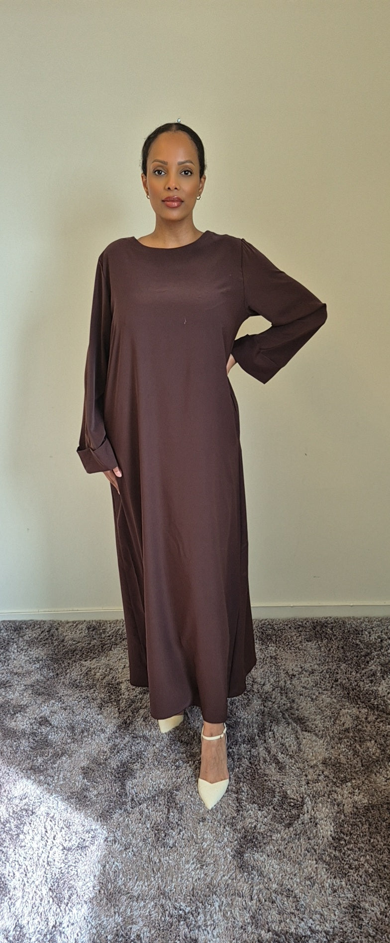 A woman poses on a gray carpet in the Elegant Black Hawra Abaya with Pockets by Bihi Boutique, featuring a long, loose fit and long sleeves, paired with beige pointed-toe heels against a plain light wall.