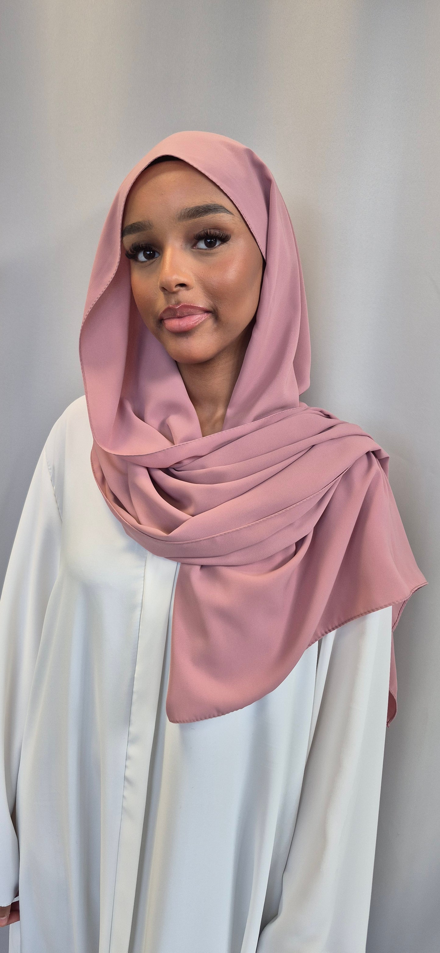 A young woman models the Medina Seide Pink Hijab by Bihi Boutique—luxurious 100% PES, 180x170 cm—paired with a white blouse, standing against a light gray background and smiling at the camera.