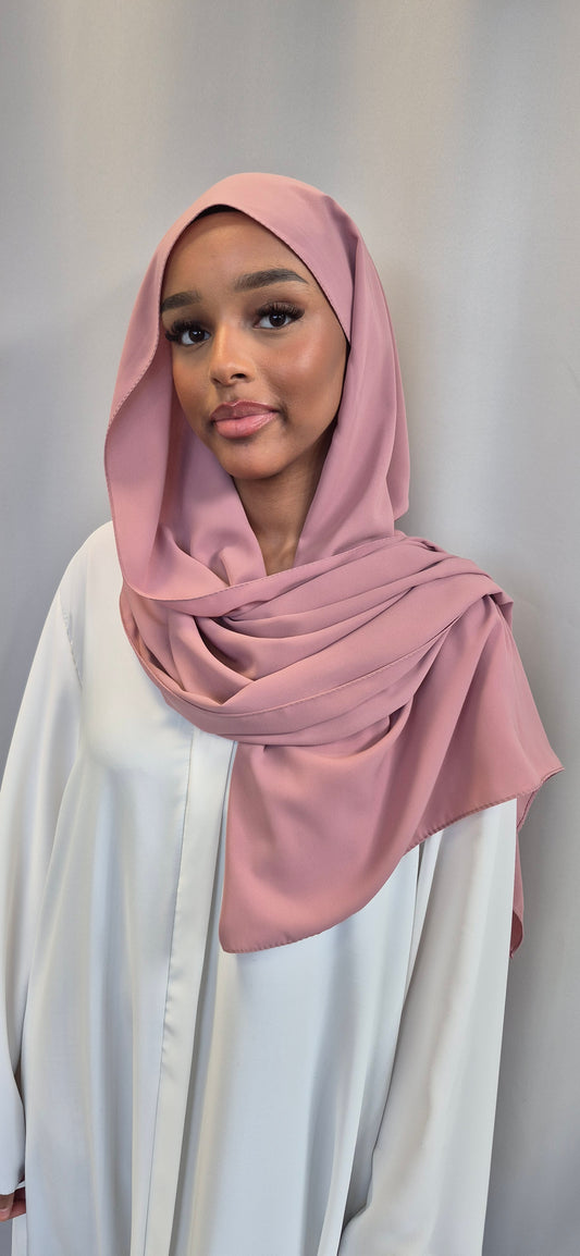 A young woman models the Medina Seide Pink Hijab by Bihi Boutique—luxurious 100% PES, 180x170 cm—paired with a white blouse, standing against a light gray background and smiling at the camera.