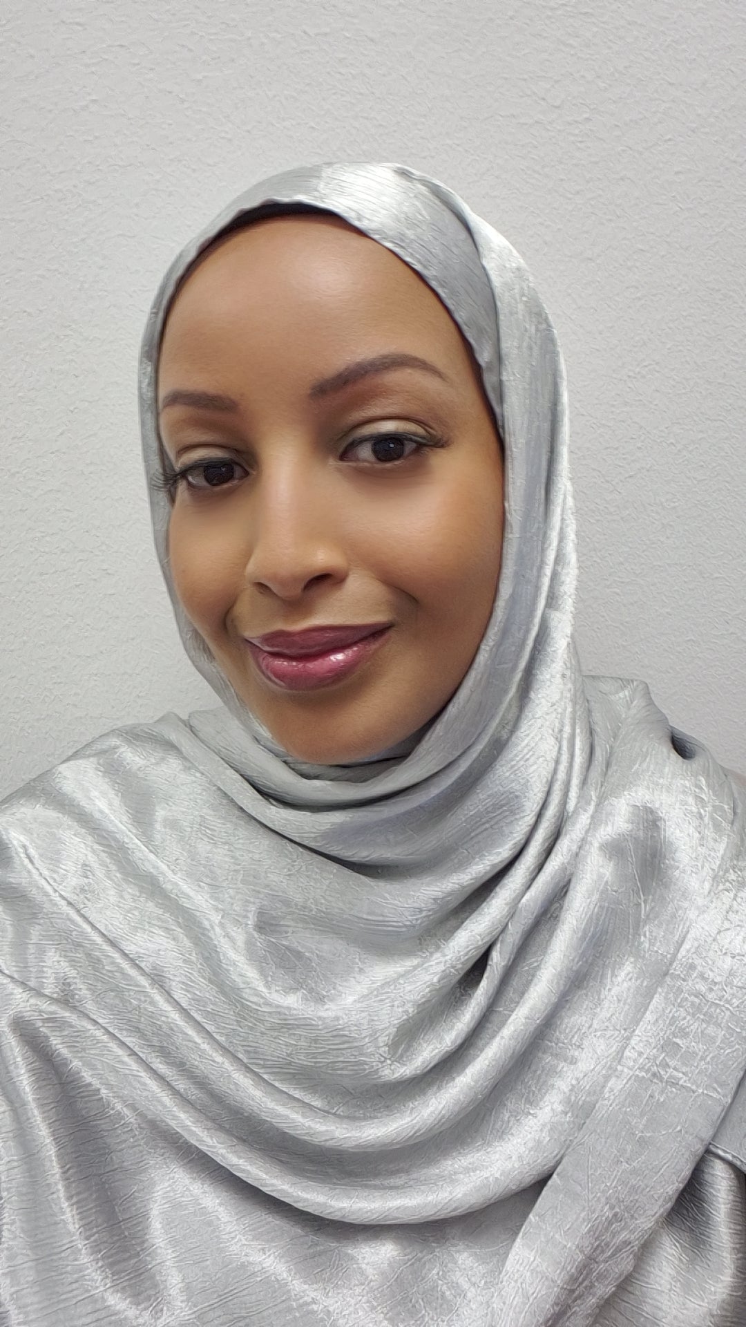 A woman wearing the Elegant Satin Hijab - Luxurious 173cm Style by Bihi Boutique smiles softly at the camera, standing against a plain light-colored wall.