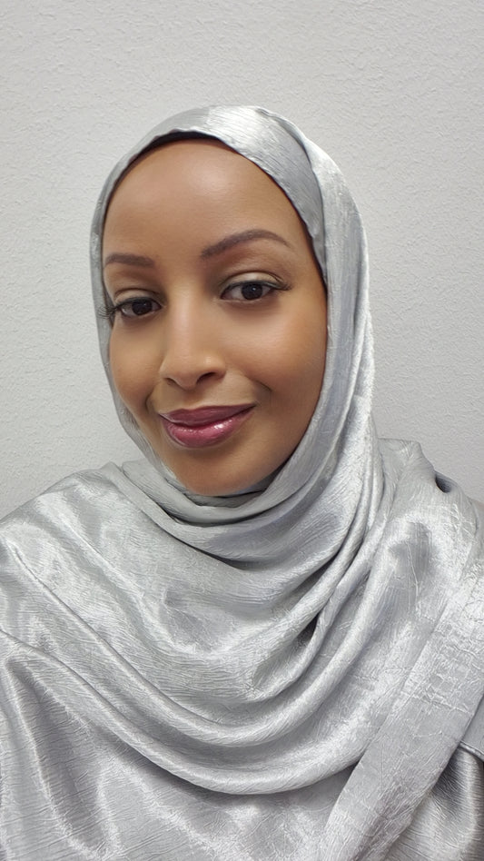 A woman wearing the Elegant Satin Hijab - Luxurious 173cm Style by Bihi Boutique smiles softly at the camera, standing against a plain light-colored wall.