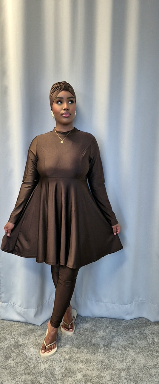 A woman poses in front of a light gray curtain, wearing the Shop Stylish Black Burkini by Bihi Boutique. She looks to the side with a neutral expression, showcasing the burkini’s perfect fit and comfort.