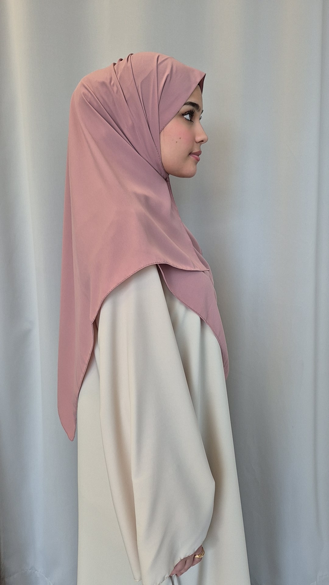 A woman in a light cream dress and a Medina Seide Pink Hijab by Bihi Boutique (100% PES, 180x170 cm) stands in profile against a plain, light-colored curtain background.