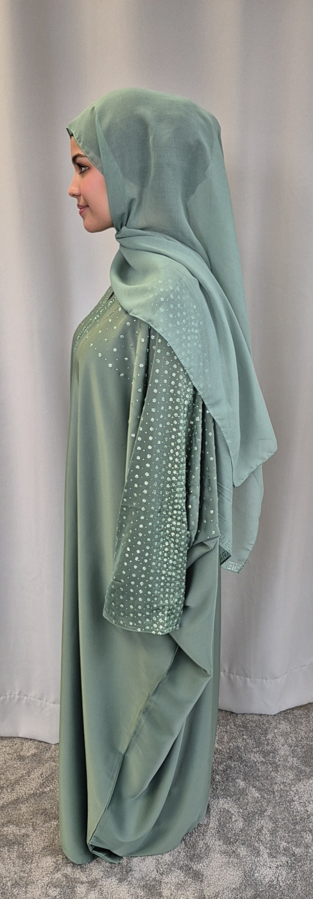 A woman in profile wears the Stylish Nourah Dress in sage green with beaded sleeves and a matching hijab by Bihi Boutique, standing before a light gray curtain and carpeted floor. Available in Perfect Fit Size 52 & 54.