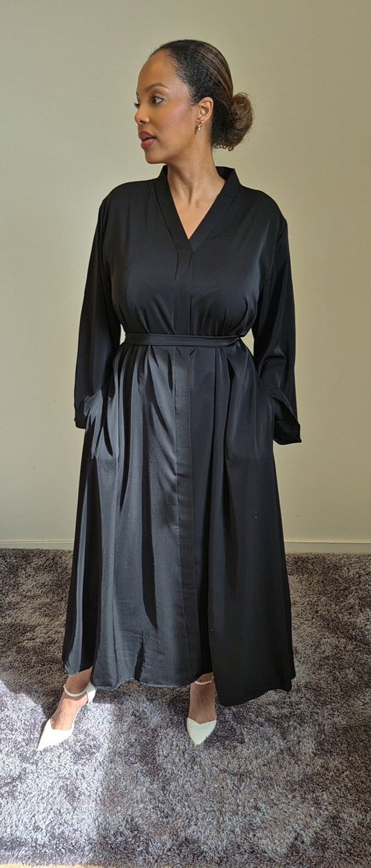 A woman stands indoors on a gray carpet wearing the Shop Layina Black Dress by Bihi Boutique in L/XL, an elegant black belted long-sleeve dress. Her hair is pulled back, she looks left, and wears white pointed shoes. Light highlights the dress.