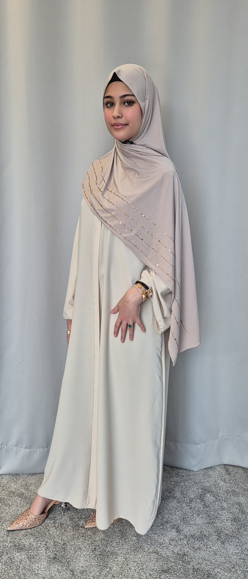 A woman in the Islene Dress by Bihi Boutique (beige, one size for 160-165 cm) and matching hijab stands before a gray curtain, styled with pointed-toe shoes and a gold watch, her makeup subtle and expression calm.