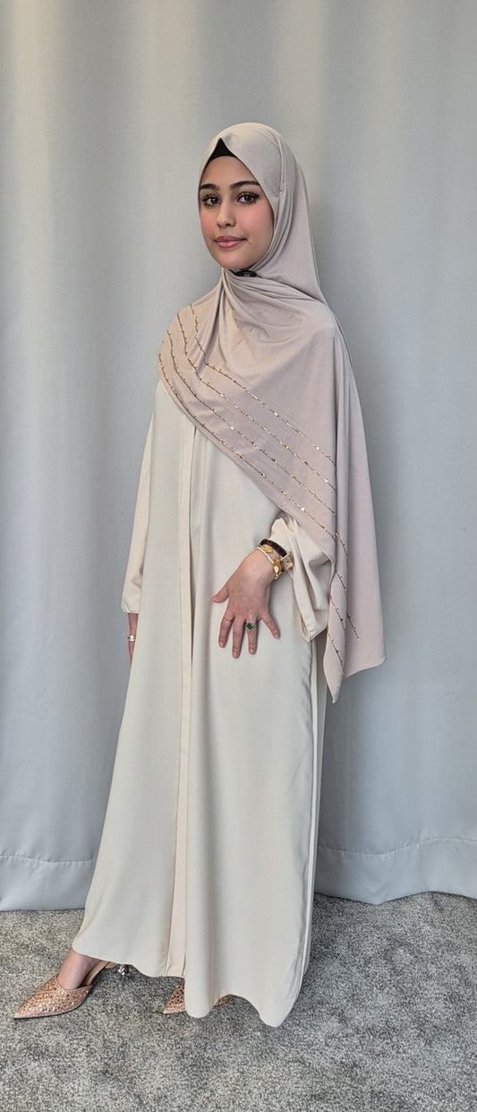A woman in the Islene Dress by Bihi Boutique (beige, one size for 160-165 cm) and matching hijab stands before a gray curtain, styled with pointed-toe shoes and a gold watch, her makeup subtle and expression calm.