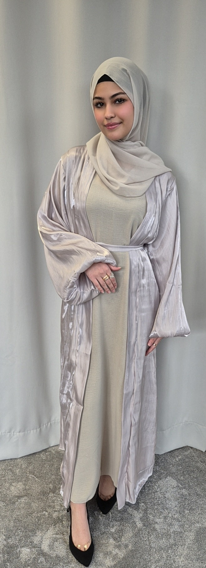 A woman dressed in the Bihi Boutique Stylish Organza Chrome Hijab Set—a chic, lightweight beige ensemble—stands on a gray carpet before a light gray curtain, smiling softly with one hand resting on her waist.