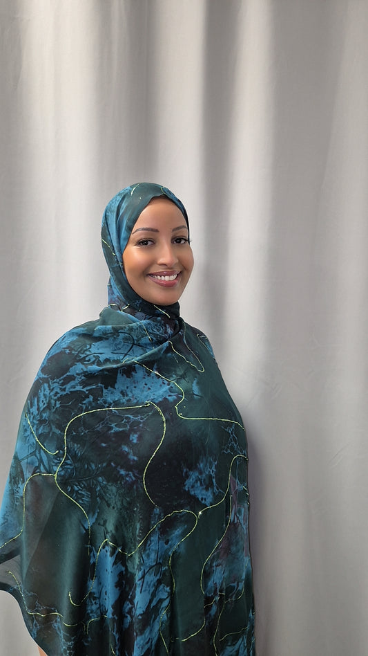 A woman wearing the Elegant Marble Chiffon Hijab from Bihi Boutique smiles warmly while standing in front of a light gray curtain.