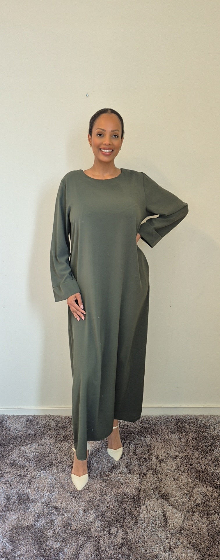 A woman stands indoors on a gray carpet, smiling at the camera while wearing the Bihi Boutique Elegant Black Hawra Abaya with Pockets. She pairs this stylish, versatile dress with white pointed-toe heels against a light-colored wall.
