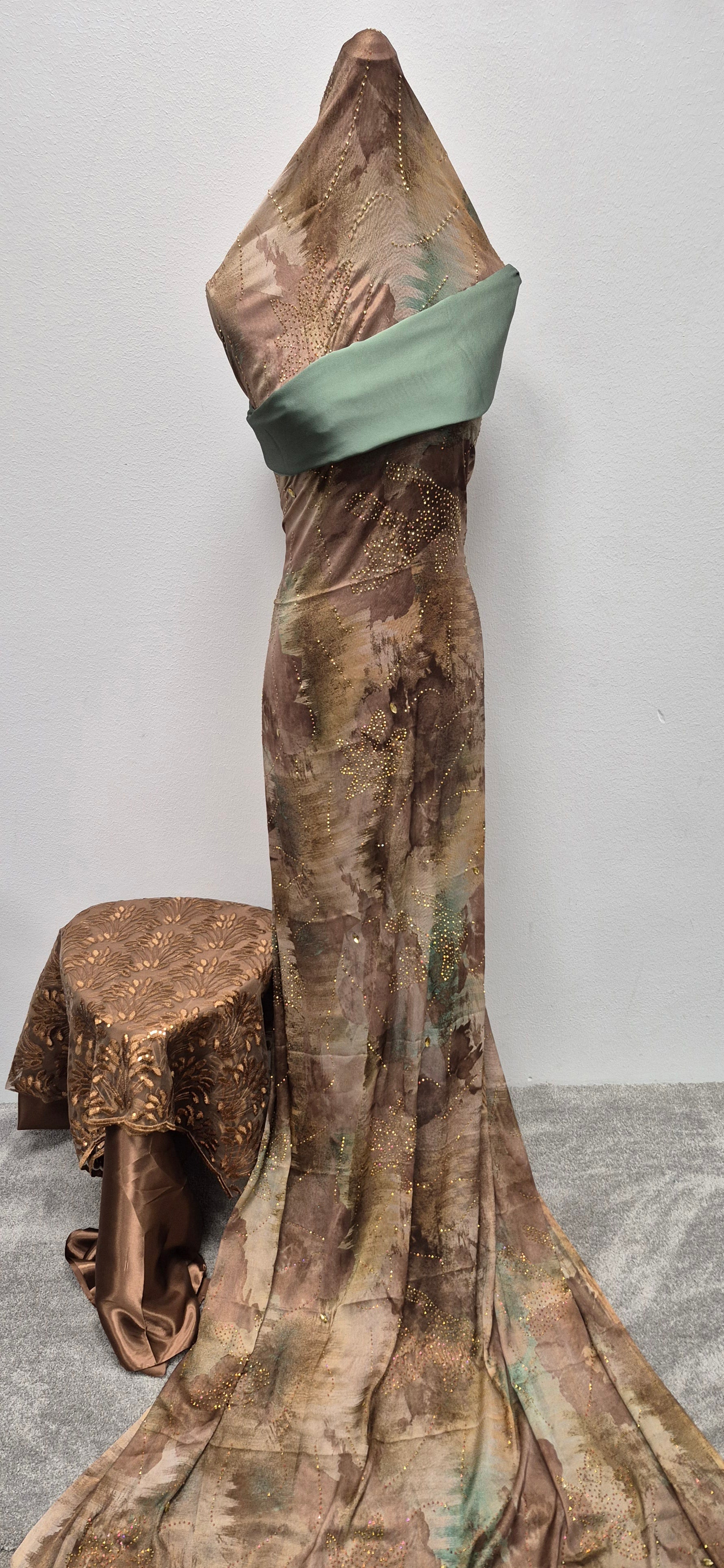 A Shop Fransawi Dirac Half Bac by Bihi Boutique, an elegant brown and gold gown with green sash, is displayed on a headless mannequin beside a matching stool on a gray carpet against a white wall.