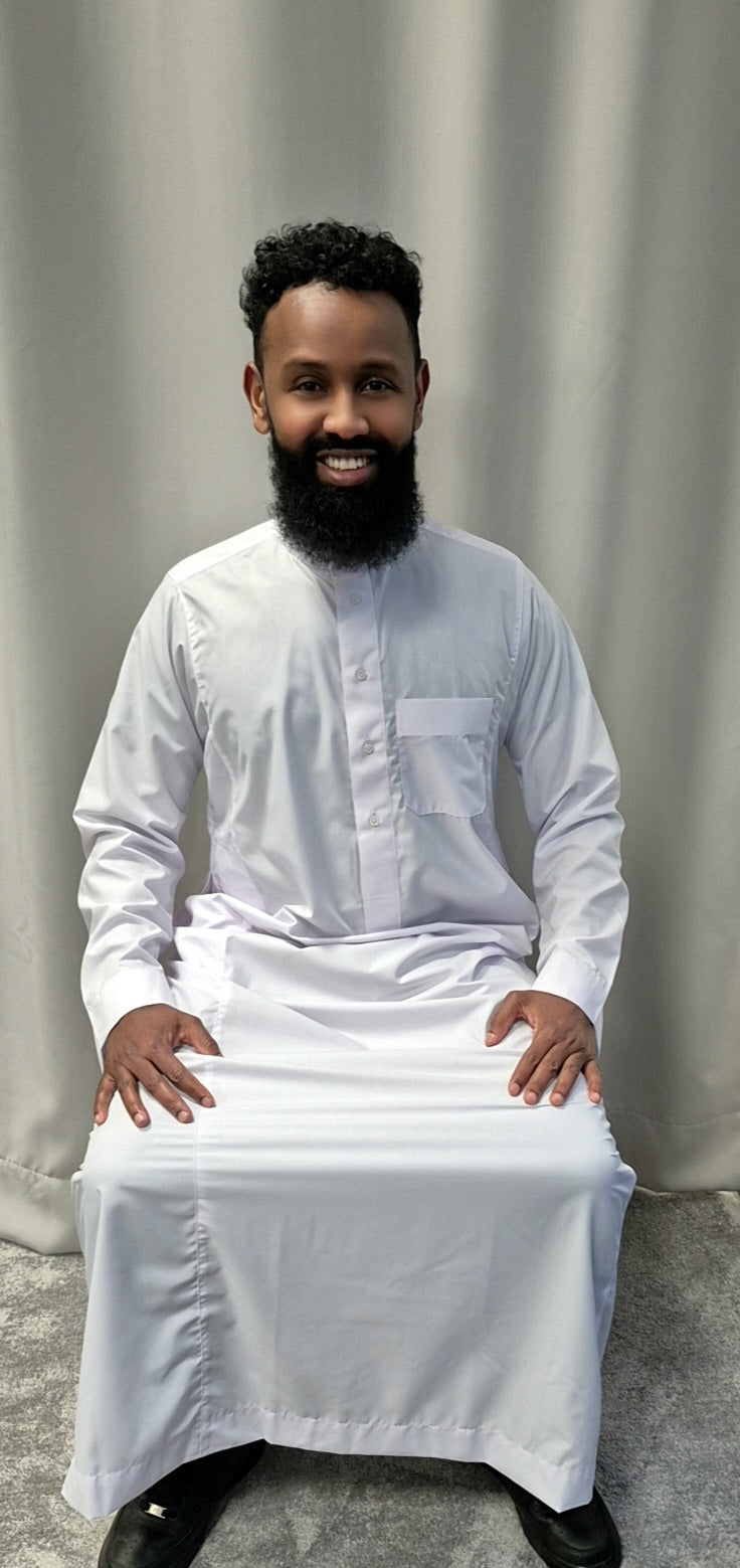 A man with a full beard and curly hair smiles at the camera, seated in front of a light gray curtain, wearing Bihi Boutiques Shop Khamis Saudi White—stylish traditional attire.