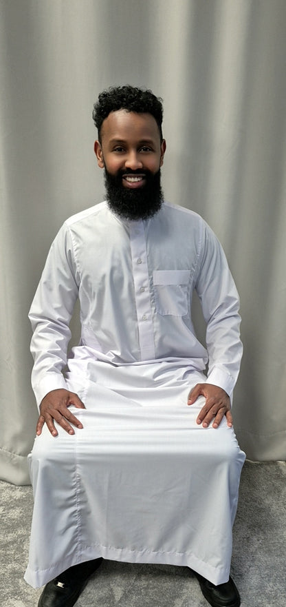 A man with a full beard and curly hair smiles at the camera, seated in front of a light gray curtain, wearing Bihi Boutiques Shop Khamis Saudi White—stylish traditional attire.