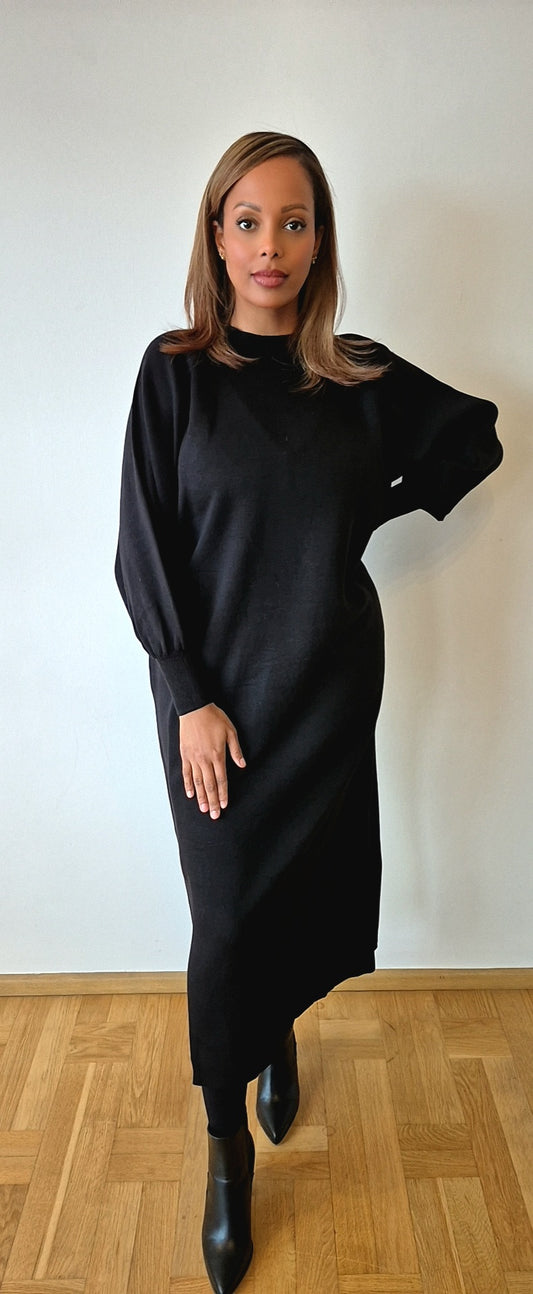 A woman stands indoors on a wooden floor, wearing the Black Modest Sweater Dress from Bihi Boutique, with black boots. She poses with one hand on her hip and the other by her side, in front of a plain white wall.