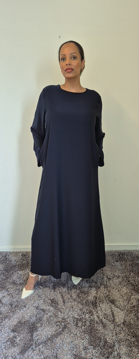 A woman stands indoors on a plush carpet, wearing the Bihi Boutique Elegant Black Hawra Abaya with Pockets. The stylish, loose-fitting abaya features long sleeves, and she faces forward with her hands in the pockets against a plain wall.