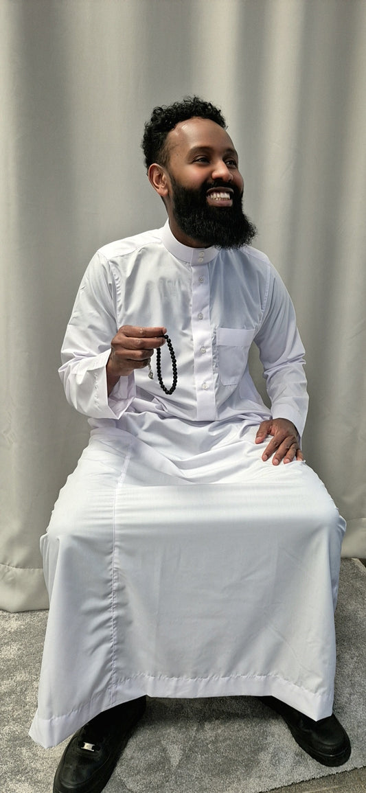 A bearded man smiles while seated, wearing the Shop Khamis Saudi White traditional attire by Bihi Boutique and holding black prayer beads in his right hand, with a light gray curtain in the background.