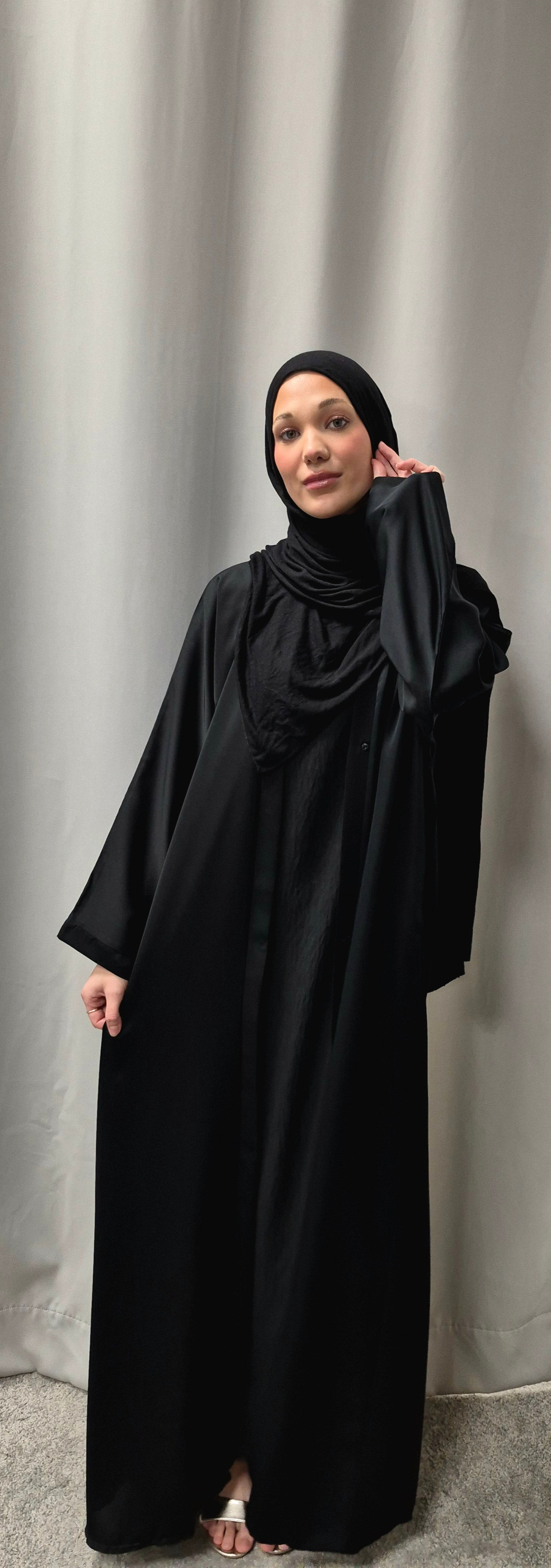 A woman models the Stylish Layla Open Abaya in Size 58 from Bihi Boutique, featuring an elegant black design and paired with a black hijab as she stands barefoot on a gray carpet before a light gray curtain.