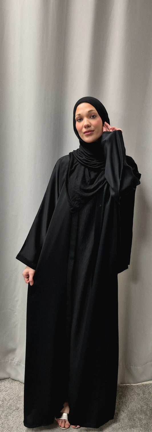 A woman models the Stylish Layla Open Abaya in Size 58 from Bihi Boutique, featuring an elegant black design and paired with a black hijab as she stands barefoot on a gray carpet before a light gray curtain.