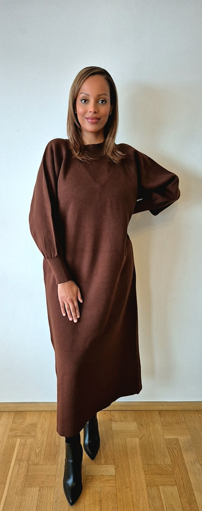 A woman with straight, shoulder-length hair poses confidently indoors against a white wall, wearing the Bihi Boutique Chic One Size Brown Sweater Dress—Cozy Wool Blend Fashion—with black ankle boots.