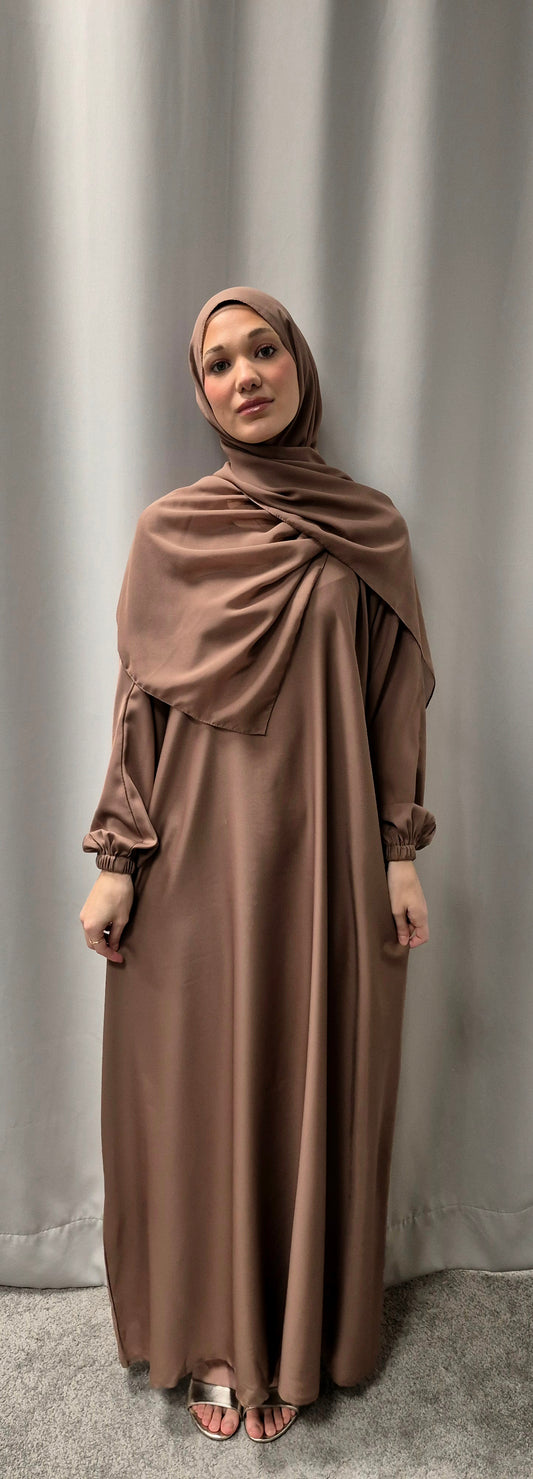 A woman stands in front of gray curtains wearing the Bihi Boutique Amina Rose Gold Abaya with a matching chiffon hijab, size 58. She looks at the camera with a neutral expression and her hands rest by her sides.