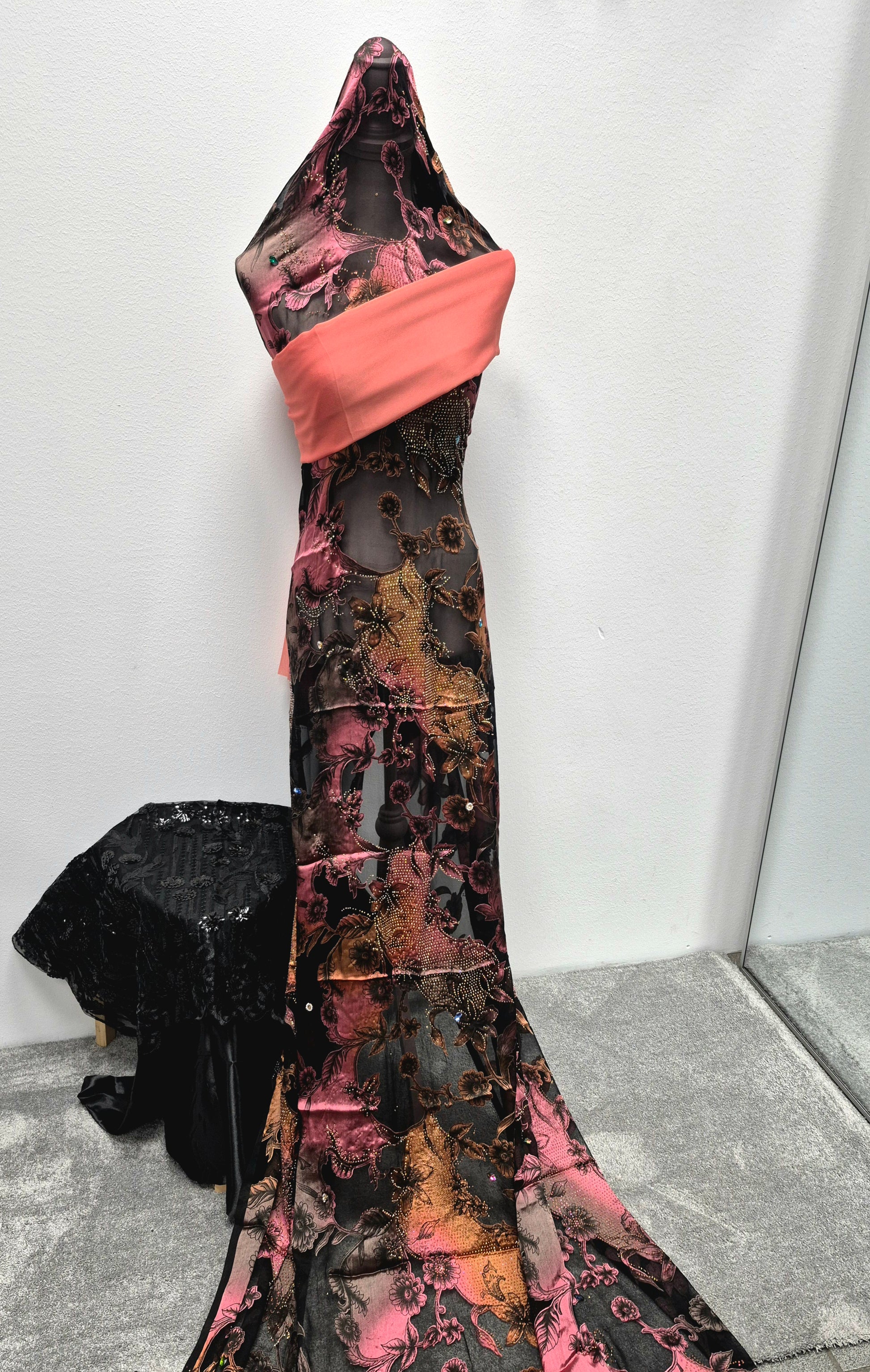 A black dress form displays the Shop Stylish Fransawi Dirac Half Bac Elegant Fashion Piece by Bihi Boutique—an elegant sheer, floor-length gown with pink and orange floral patterns and a coral sash, shown on gray carpet near a mirror and draped black lace.