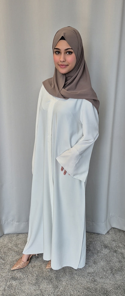 A woman in the Bihi Boutique Islene Dress (white, one size fits 160-165 cm) and a taupe hijab stands on a gray carpet before a light gray curtain, smiling slightly with one hand by her side.