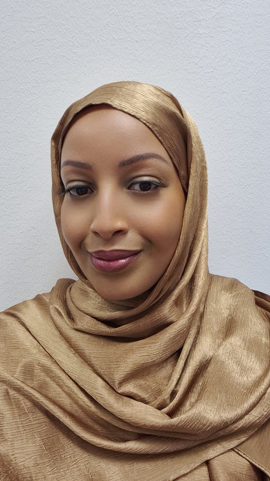 A woman smiles softly at the camera, wearing Bihi Boutique’s Stylish Satin Hijab (173cm x 73cm) in a shiny gold color, set against a plain white background.